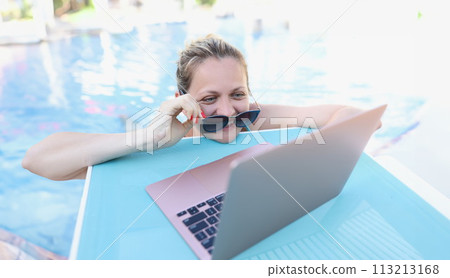 Woman straightening sunglasses and looking into laptop screen in swimming pool 113213168
