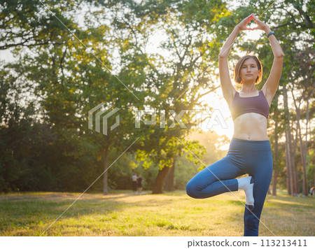 Young asian woman doing yoga in morning or evening at park, healthy woman relaxing and practicing tree pose at city park. Mindfulness, destress, Healthy habits and balance concept 113213411