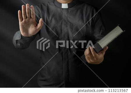 Young priest holding bible and raised hand giving forgiveness isolated on black background. 113213567