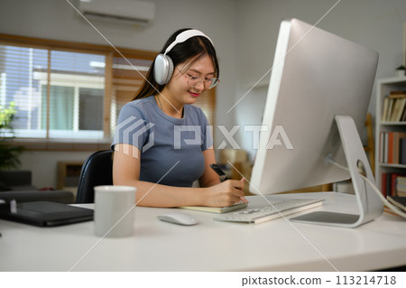 Young woman in headphone working at home, making notes on notepad Young woman in headphone working at home, making notes on notepad 113214718