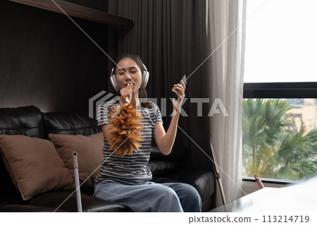 Carefree young woman in headphones cleaning house and singing, using duster as microphone 113214719
