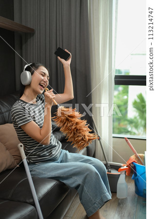 Overjoyed young housewife in headphones singing while cleaning living room 113214747