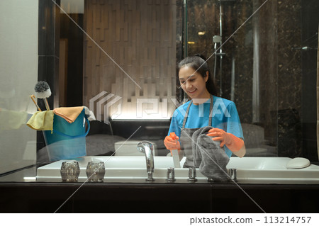 Smiling housekeeper in rubber gloves washing bathtub and faucet in a modern bathroom Smiling housekeeper in rubber gloves washing bathtub and faucet in a modern bathroom 113214757