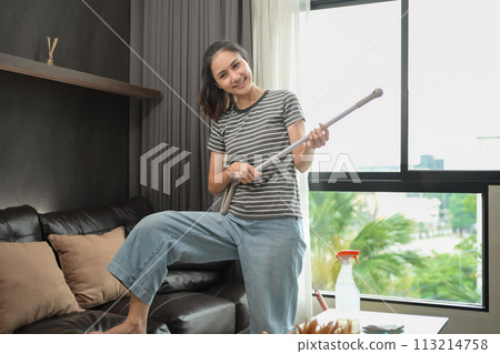 Cheerful young woman pretending to play guitar with broom while cleaning house Cheerful young woman pretending to play guitar with broom while cleaning house 113214758