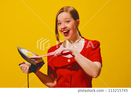 Smiling young woman in red dress cooking egg on iron, laughing against yellow background. Creative poster for cooking school and culinary workshop. 113215745