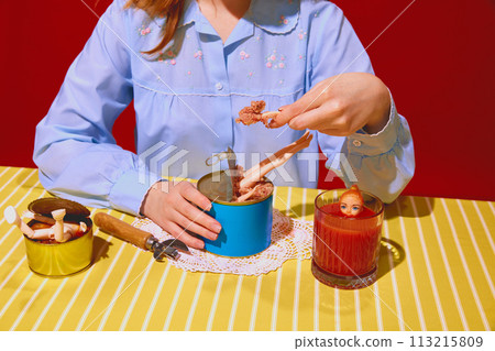 Young woman in blue blouse with doll parts on meat can, drinking tomato juice against red background Young woman in blue blouse with doll parts on meat can, drinking tomato juice against red background 113215809
