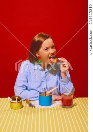 Young woman sitting at table with cans and eating doll with tomato juice against red background. Quirky dining Young woman sitting at table with cans and eating doll with tomato juice against red background. Quirky dining 113215810