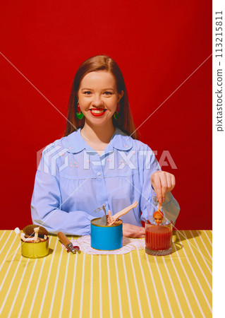 Smiling young woman sitting at table with cans and putting doll inside tomato juice against red background. Quirky dining 113215811
