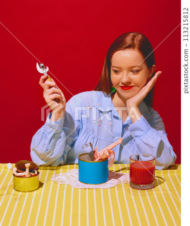 Smiling young woman holding can opener with cans and doll parts inside, tomato juice against red background. Unique and eccentric dining experiences Smiling young woman holding can opener with cans and doll parts inside, tomato juice against red background. Unique and eccentric dining experiences 113215812