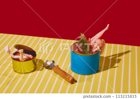 Doll parts in tin cans with can opener on striped tablecloth against red background. Doll parts in tin cans with can opener on striped tablecloth against red background. 113215815