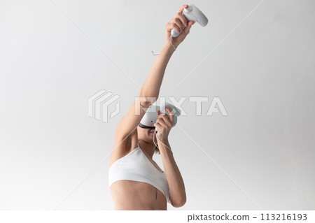 Beautiful thin girl in white shorts and a top in a virtual reality helmet posing on a white background 113216193