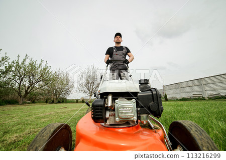 Professional gardener in protective apparel is mowing green grass lawn using modern gasoline cordless lawnmower at backyard. Seasonal landscaping design work. Blooming trees on background Professional gardener in protective apparel is mowing green grass lawn using modern gasoline cordless lawnmower at backyard. Seasonal landscaping design work. Blooming trees on background 113216299