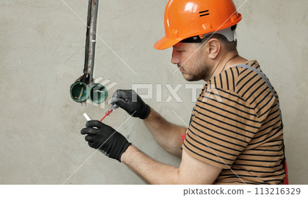 Electrician male in uniform, protective gloves and helmet checks presence of electrical voltage in socket phase uses electrical tester screwdriver. Examining wires in outlet by voltage detector indoor 113216329