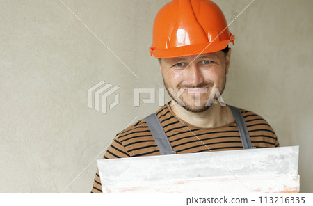 Smiling handyman repairman worker in overalls and protective orange helmet holds big putty knife for plastering concrete wall with putty. Renovation at apartment and construction new building concept. 113216335