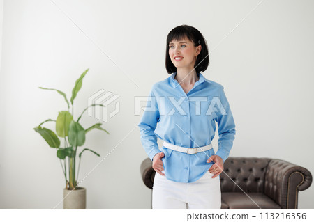Women equality day. Smiling successful businesswoman in elegant blue shirt and white pants looks at camera at modern comfortable light office with green plant and sofa. Work life balance concept. Women equality day. Smiling successful businesswoman in elegant blue shirt and white pants looks at camera at modern comfortable light office with green plant and sofa. Work life balance concept. 113216356
