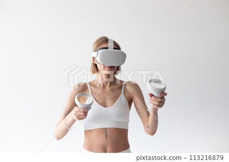 Beautiful thin girl in white shorts and a top in a virtual reality helmet posing on a white background 113216879