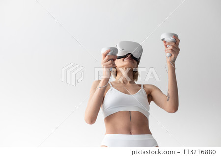 Beautiful thin girl in white shorts and a top in a virtual reality helmet posing on a white background 113216880