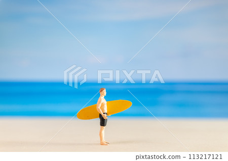 Miniature people man in a swimsuit, and holding a yellow surfboard on the beach 113217121