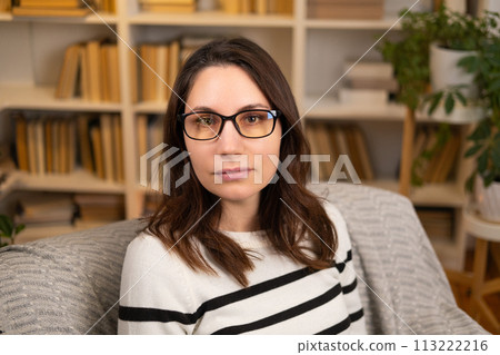 Portrait of a young adult beautiful woman in glasses and a striped blouse sitting in an sofa and looking at the camera indoors 113222216