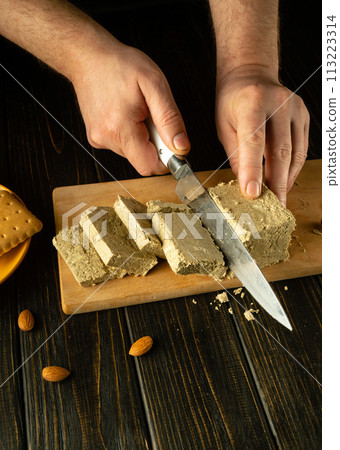 The cook uses a knife to cut sunflower halva into slices. Low key concept of serving a sweet and delicious dessert for breakfast with tea. Space for recipe or menu 113223314