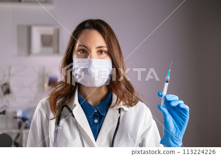 A young adult female doctor in a medical mask and white coat holds a syringe in her hands and looks at the camera 113224226