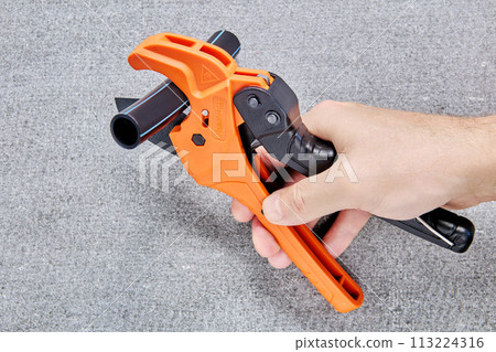 Ratcheting pipe cutter in shape of scissors with polyethylene tube on blade is held in hand of plumber. Ratcheting pipe cutter in shape of scissors with polyethylene tube on blade is held in hand of plumber. 113224316