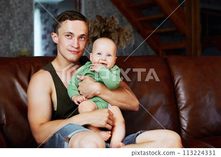 Caucasian man, 26 years old, holds his 6 month old baby daughter in his arms, sitting at home on leather sofa in living room. Caucasian man, 26 years old, holds his 6 month old baby daughter in his arms, sitting at home on leather sofa in living room. 113224331