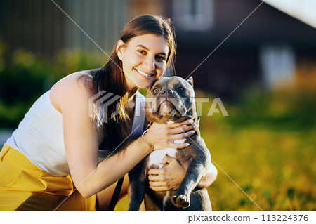 Young woman raised muzzle of American Bully to take joint portrait with her dog, white female is photographed with her pet on summer evening on lawn. 113224376