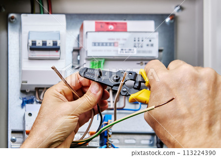 An electrician uses stripper cutter tool to remove insulation from ends of wires when connecting power cable to electrical network through an outdoor distribution panel. 113224390