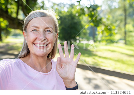 Close-up portrait of a smiling senior gray-haired woman standing outside in a park, talking and greeting on a mobile phone camera. Close-up portrait of a smiling senior gray-haired woman standing outside in a park, talking and greeting on a mobile phone camera. 113224631