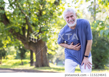 An elderly gray-haired man walks in the park and does sports, stands bent and twisted in pain, holding his stomach with his hand. 113224641