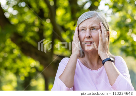 Close-up photo of an older gray-haired woman standing outside in the park and holding her head with her hands, feeling severe pain and pressure, giving a massage. 113224648