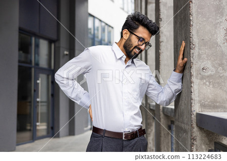 Indian young man suffering from fatigue and back pain, standing against the office wall outside and holding his lower back. 113224683