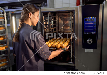 Young woman baker in uniform baking fresh baguettes in the bakery 113225241