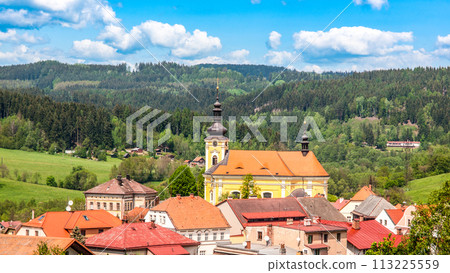 The Church of Saint Bartholomew in Pecka towers over quaint houses amidst lush green hills under a clear blue sky. Czechia 113225559