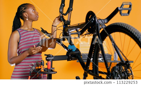 African american woman doing bike servicing in studio background repair shop, looking on tablet maintenance list. Technician checking bicycle components that need repairing, camera B African american woman doing bike servicing in studio background repair shop, looking on tablet maintenance list. Technician checking bicycle components that need repairing, camera B 113229253