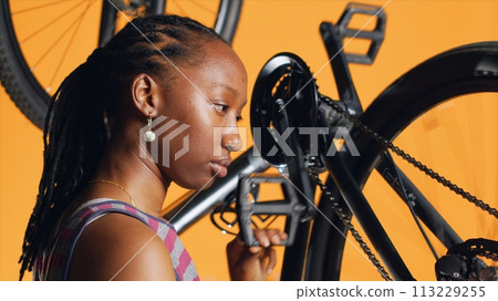 Smiling african american woman satisfied while pushing pedals to rotate bike wheel, after managing to repair it. Professional pleased with results after adjusting defective bike crank arm 113229255