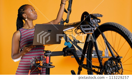 Technician with laptop in hand analyzing broken bike over studio background awaiting repair. Specialist using notebook to look for replacement parts for bicycle on repair stand, camera B Technician with laptop in hand analyzing broken bike over studio background awaiting repair. Specialist using notebook to look for replacement parts for bicycle on repair stand, camera B 113229257