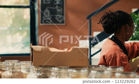 Vendor placing crates of fruits and veggies on shelves after organic produce procurement in local supermarket. African american woman arranging natural eco friendly merchandise. Tripod shot. 113229262