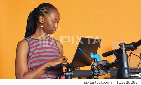 Cheerful mechanic doing bike servicing in studio background atelier shop, looking on laptop maintenance list. Specialist checking online for replacement bicycle parts using notebook, camera A 113229265