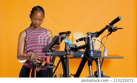 Woman following internet tutorial clips on tablet, learning to fix her damaged bike. Cycling hobbyist following guide on how to do upkeeping for bicycle components, studio background, camera B 113229272