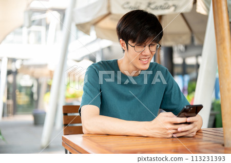 A smiling young Asian man wearing glasses sits at a wooden table outdoors, looking at his smartphone A smiling young Asian man wearing glasses sits at a wooden table outdoors, looking at his smartphone 113231393