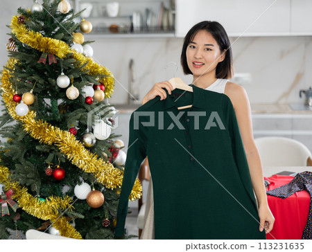 Happy young Asian girl trying to choose a dress standing next to Xmas-tree 113231635