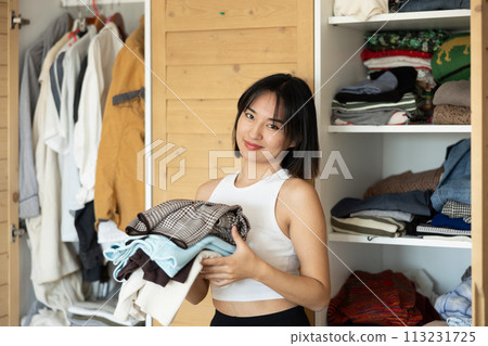 Happy young Asian girl holding some items of clothing by the wardrobe Happy young Asian girl holding some items of clothing by the wardrobe 113231725