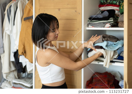 Happy young Asian girl putting some items of clothing in the wardrobe Happy young Asian girl putting some items of clothing in the wardrobe 113231727