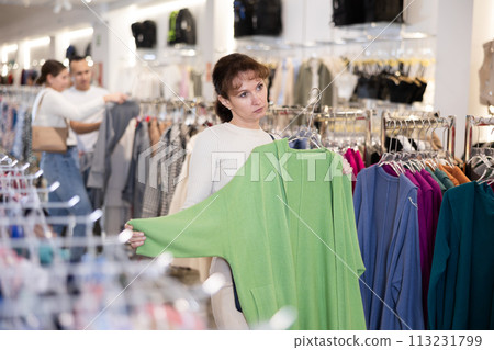 Woman stopped near clothing showcase, holds cardigan hanger in hands, examines product Woman stopped near clothing showcase, holds cardigan hanger in hands, examines product 113231799