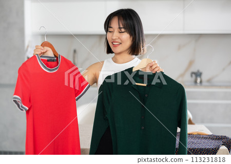 Happy young Asian girl trying to choose a dress in the kitchen Happy young Asian girl trying to choose a dress in the kitchen 113231818