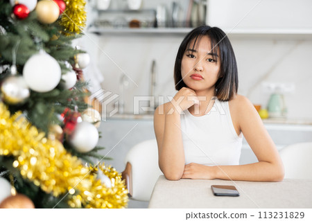 Sad young Asian girl waiting for phone call sitting at table by Xmas tree Sad young Asian girl waiting for phone call sitting at table by Xmas tree 113231829