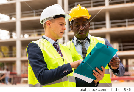 Two civil engineers are discussing the construction plan, holding documents and studying them 113231986