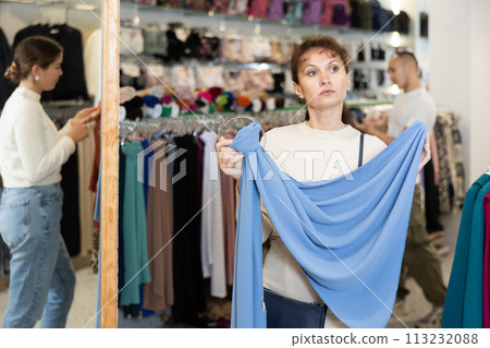 Woman stopped near accessories showcase, holds silk scarf hanger in hands, examines product Woman stopped near accessories showcase, holds silk scarf hanger in hands, examines product 113232088